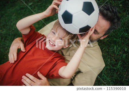 Dad lying on grass with son, laughing and having fun. Father and boy playing football, catching each other, falling to the ground. Fatherhood and Father's Day. 115390010