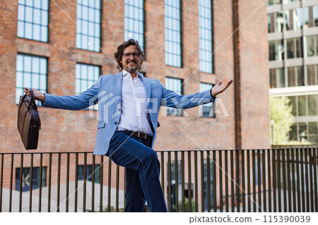 Portrait of happy mature businessman standing on city street, in front of office building. Handsome man with glasses in suit celebrating successful business deal. 115390039