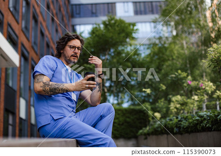 Healthcare worker taking a break, smoking a cigarette outdoors and scrolling on phone, in front of the hospital building. 115390073
