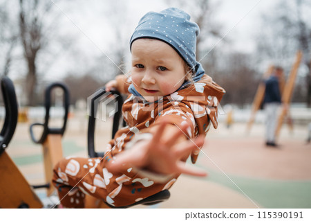 Cute toddler girl playing outdoor in public park playground. Girl in softshell bodysuit spending time in park. Cute toddler girl playing outdoor in public park playground. Girl in softshell bodysuit spending time in park. 115390191
