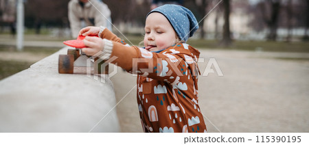 Cute toddler girl playing outdoor in playground with skateboard. Girl in softshell bodysuit spending time in park. Cute toddler girl playing outdoor in playground with skateboard. Girl in softshell bodysuit spending time in park. 115390195