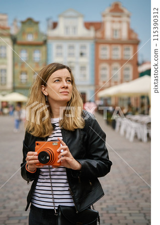 Woman taking picture using orange instant camera. Tourist captures memories during travel with vintage camera. Female walking around city street 115390312