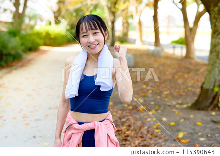 A young woman jogging along a road lined with trees in an autumn park, fist pump, diet, body makeover, towel in hand A young woman jogging along a road lined with trees in an autumn park, fist pump, diet, body makeover, towel in hand 115390643
