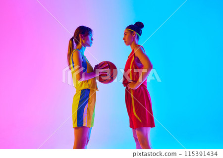 Two women, basketball players in uniforms, opponents standing and looking at each other with focus against gradient pink blue background in neon light. 115391434