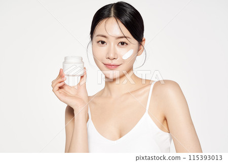 an Asian woman with cream on her face and holding cosmetics 115393013
