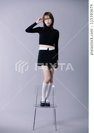 A Korean woman in her 20s posing to touch glasses while standing on a chair wearing a geek look A Korean woman in her 20s posing to touch glasses while standing on a chair wearing a geek look 115393674