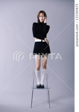 A Korean woman in her 20s posing to touch glasses while standing on a chair wearing a geek look 115393675