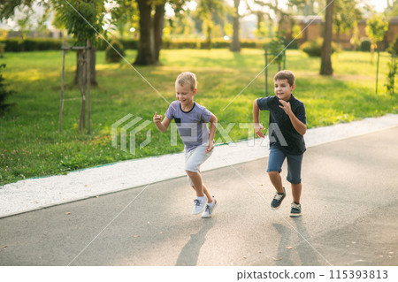 Two children are playing in the park. Two beautiful boys in T-shirts and shorts have fun smiling. They eat ice cream, jump, run. Summer is sunny Two children are playing in the park. Two beautiful boys in T-shirts and shorts have fun smiling. They eat ice cream, jump, run. Summer is sunny 115393813