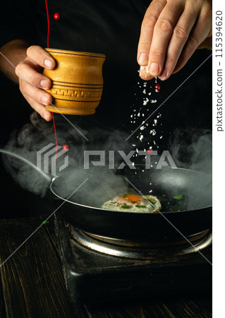 The chef manually adds salt to a hot pan of eggs. Low key concept of preparing breakfast with egg in restaurant kitchen The chef manually adds salt to a hot pan of eggs. Low key concept of preparing breakfast with egg in restaurant kitchen 115394620