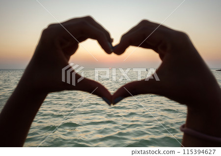 Close up of woman hands makes a heart shape against the sunset sky and sea Close up of woman hands makes a heart shape against the sunset sky and sea 115394627
