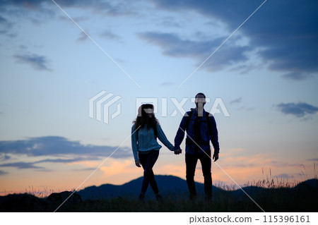 Travellers couple hiking on trail in nature with backpacks, during sunset. Young tourist spending summer vacation oudoors, holding hands. 115396161