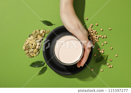 Cup of milk in hand, leaves, soybean and pumpkin seeds on green background, top view Cup of milk in hand, leaves, soybean and pumpkin seeds on green background, top view 115397277