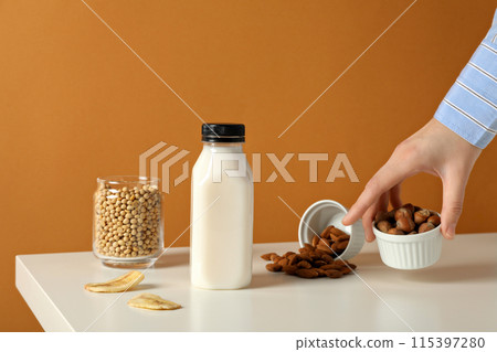 Bottle of milk, hand, nuts in bowls, slices of banana and soy in glass on orange background 115397280