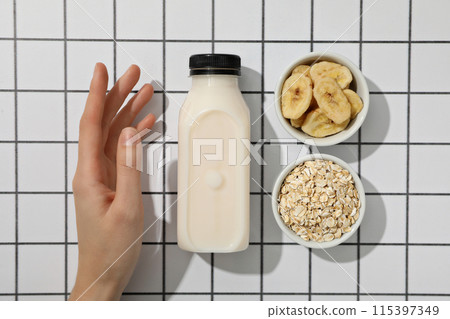 Bottle with milk, hand and bowls with oatmeal and sliced bananas on light background, top view 115397349