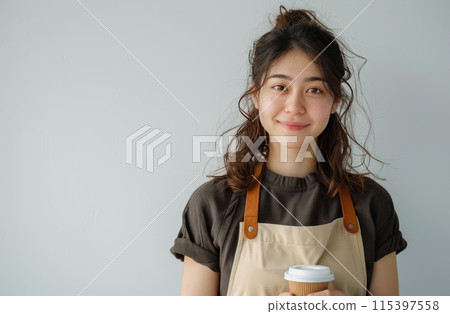 Young Woman With Brown Hair Wearing Apron Holds Coffee Cup Against White Wall 115397558