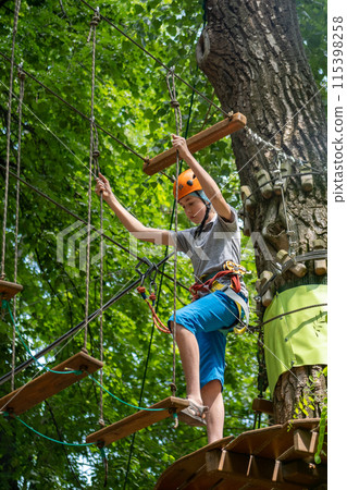 Rope park. A boy teenager in a helmet walks on suspended rope ladders. Carabiners and safety straps. Safety. Summer activity. Sport. Children's playground in nature in the forest 115398258