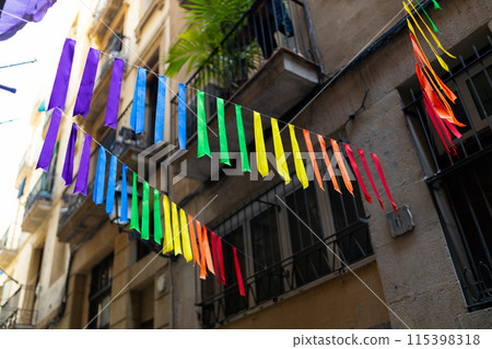 Festive ribbon of multicolored rainbow flags on the urban background. Celebration party, holiday fair, festival decorations. Gay pride symbol. Festive ribbon of multicolored rainbow flags on the urban background. Celebration party, holiday fair, festival decorations. Gay pride symbol. 115398318