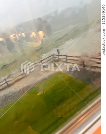A view through a rain-streaked window capturing a heavy downpour with hail and flash flooding in a suburban backyard. The scene shows blurred fences, trees, and lawn due to the intense rain and hail. 115399146