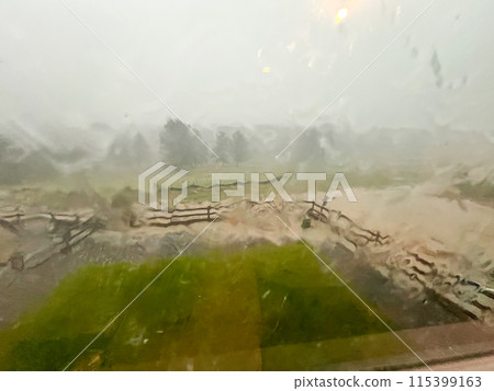 A view through a rain-streaked window capturing a heavy downpour with hail and flash flooding in a suburban backyard. The scene shows blurred fences, trees, and lawn due to the intense rain and hail. 115399163