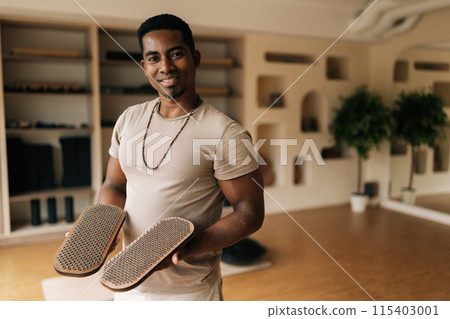 Portrait of cheerful African man holding in hands wooden Sadhu Board with nails standing in cozy living room, preparing to practice, smiling looking at camera. Concept of mindfulness, tranquility. 115403001