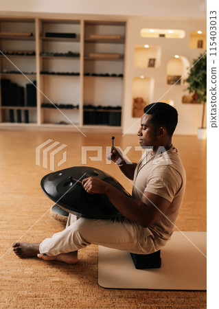 Vertical side view of serene black male playing ethnic native music on tank drum with sticks sitting cross-legged on floor home. African drummer using traditional meditative instrument glucophone. 115403013