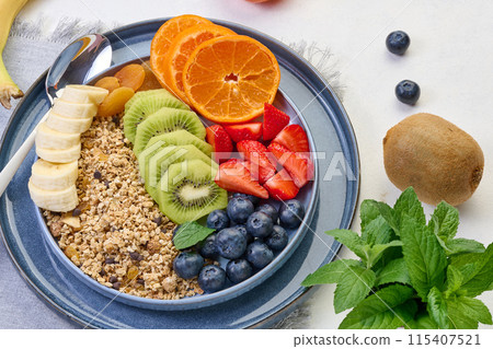 Granola with strawberries, kiwi, banana and blueberries in a round plate on a white table. 115407521