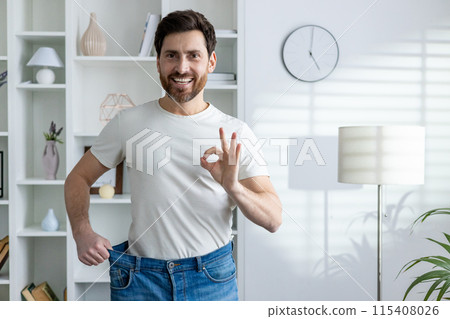 Young smiling and happy man standing at home in oversized jeans and showing his result of weight loss and dieting, showing ok sign with his hand. 115408026