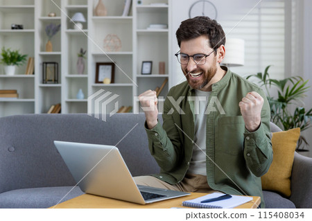 A happy young man sits at home on the sofa in front of a laptop and rejoices in success, showing a victory gesture with his hands, looks contentedly at the screen, rejoices at the received news. 115408034