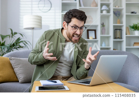 Angry young man in glasses and green shirt sitting on sofa at home in front of laptop and gesturing emotionally at screen, got bad news, talking on video call. 115408035