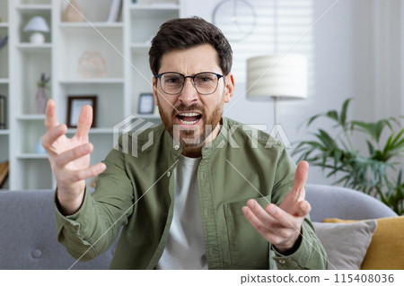 Close-up portrait of angry and worried young man sitting on sofa at home, talking on video call and emotionally shouting at camera, gesturing with hands. 115408036