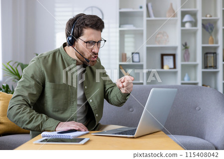 Annoyed young man in headset sitting on sofa at home and talking on video call via laptop, emotionally arguing pointing finger at monitor. 115408042