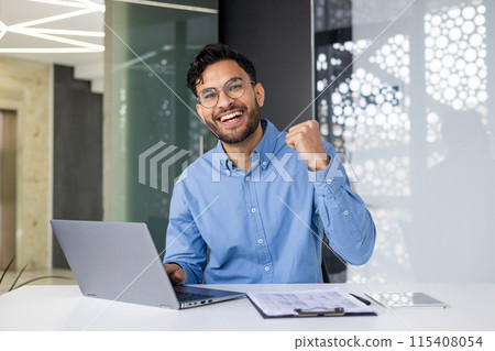 Smiling business professional celebrating success while working on a laptop in a modern office environment. The person is feeling happy and accomplished. 115408054