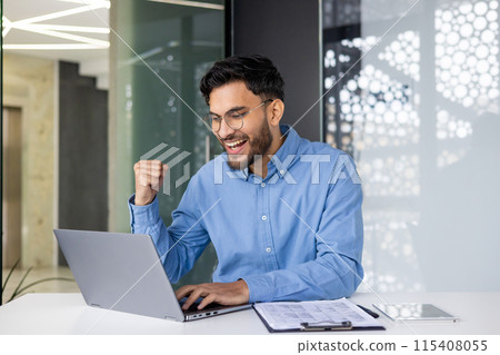 Happy young Indian man working in the office, sitting with a chair and enjoying success, looking at the laptop screen and showing a victory gesture with his hand. 115408055