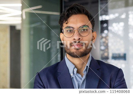 Close-up portrait of a serious young Indian man in a business suit and glasses in the office, looking confidently at the camera. 115408068