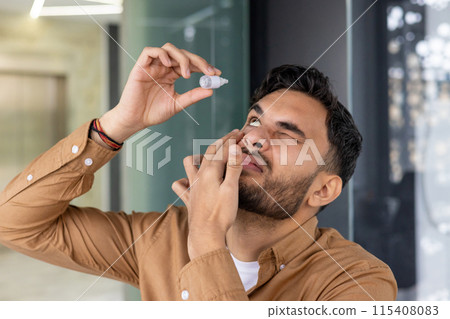 Close-up photo of a young Indian man standing in a modern office and instilling medicine for fatigue and inflammation into his eye. Close-up photo of a young Indian man standing in a modern office and instilling medicine for fatigue and inflammation into his eye. 115408083