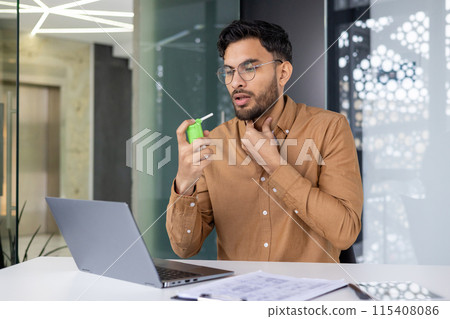 Young businessman using a throat spray while working on a laptop in a modern office. He appears to be dealing with a sore throat. 115408086
