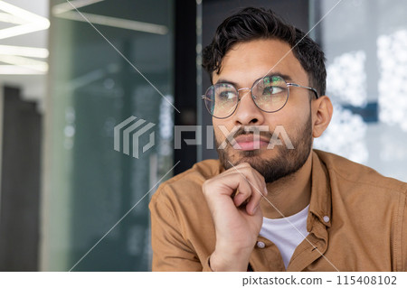 Close-up of a thoughtful young man wearing glasses and looking away, contemplating an idea while sitting indoors. 115408102