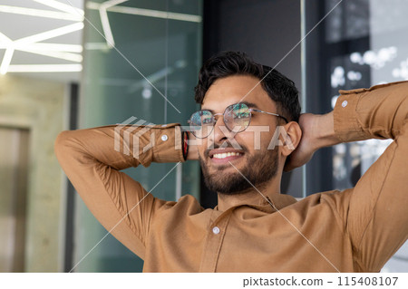 Relaxed young man with glasses smiling confidently in a modern office setting. Brown shirt, casual pose, and contemporary background. Relaxed young man with glasses smiling confidently in a modern office setting. Brown shirt, casual pose, and contemporary background. 115408107