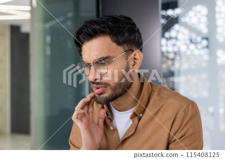 A young man with glasses is seen experiencing a toothache while sitting in a modern office environment. 115408125
