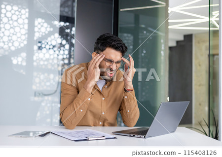A tired young Indian man sits in the office at the desk and hides from the pain and holds his head with his hands, feels a lot of pressure. A tired young Indian man sits in the office at the desk and hides from the pain and holds his head with his hands, feels a lot of pressure. 115408126