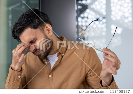 Young man experiencing eye strain while removing his eyeglasses in a modern office setting, conveying discomfort, stress, and fatigue. Young man experiencing eye strain while removing his eyeglasses in a modern office setting, conveying discomfort, stress, and fatigue. 115408127