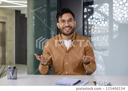 Smiling young Indian man sitting in the office at the desk and talking on a video call, making an online recording, conducting training remotely. Smiling young Indian man sitting in the office at the desk and talking on a video call, making an online recording, conducting training remotely. 115408134
