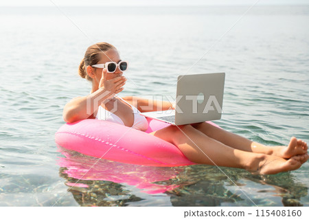 A woman is sitting on a pink inflatable raft in the ocean, using a laptop. Concept of relaxation and leisure, as the woman is enjoying her time in the water while working on her laptop. 115408160