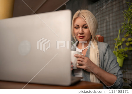 A blonde woman is sitting at a table with a laptop and a white coffee cup. She is wearing a gray jacket and has red lipstick on. The scene suggests a casual and relaxed atmosphere. A blonde woman is sitting at a table with a laptop and a white coffee cup. She is wearing a gray jacket and has red lipstick on. The scene suggests a casual and relaxed atmosphere. 115408171