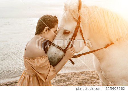 A woman in a dress stands next to a white horse on a beach, with the blue sky and sea in the background. A woman in a dress stands next to a white horse on a beach, with the blue sky and sea in the background. 115408262