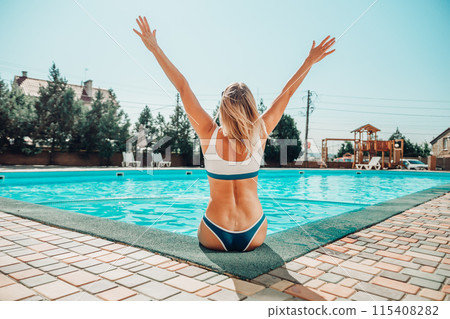 A woman is sitting in a pool with her arms raised in the air. Scene is joyful and carefree. 115408282