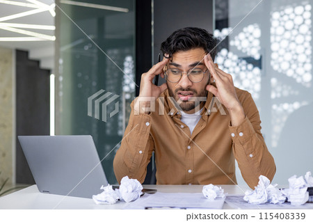 Shocked and upset young Indian man sitting at desk with laptop, crumpled and thrown away paper, tired and frustrated holding his head looking for new ideas. Shocked and upset young Indian man sitting at desk with laptop, crumpled and thrown away paper, tired and frustrated holding his head looking for new ideas. 115408339