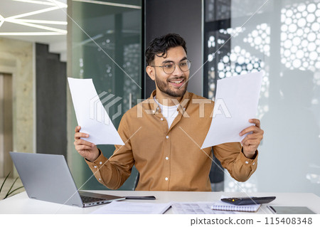 Smiling businessman holds papers while working on a laptop at his office desk. He's reviewing documents and feeling positive. 115408348