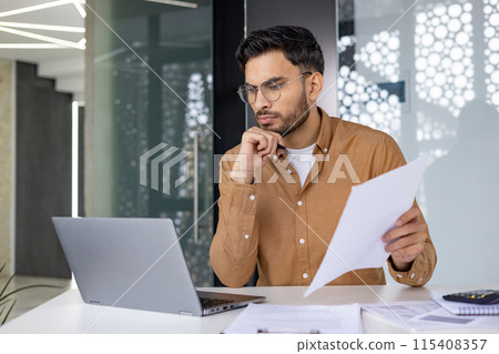 Focused businessman wearing glasses, working on his laptop while holding documents in a modern, well-lit office. Concentration and productivity in a professional workspace. 115408357