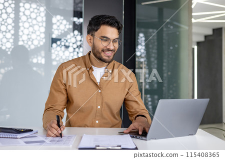 Smiling Indian man working on a laptop in the office, sitting at a table in front of a screen, writing documents and talking on a video call. 115408365